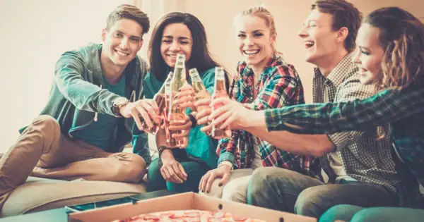 Grupa przyjaciół wznosi toast butelkami piwa, śmiejąc się i jedząc pizzę. Nawet trudne pytania nie psują im nastroju.